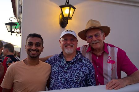 Three men smiling and posing together at an outdoor gathering near a white wall with lantern lights.