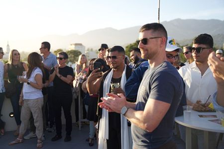 A group of people outdoors clapping and smiling during a rooftop event with mountains in the background.