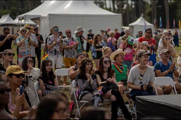 A diverse crowd of attendees watches a stage performance at the Pacific Pride Festival, seated in rows of white chairs with tents and rainbow flags in the background.