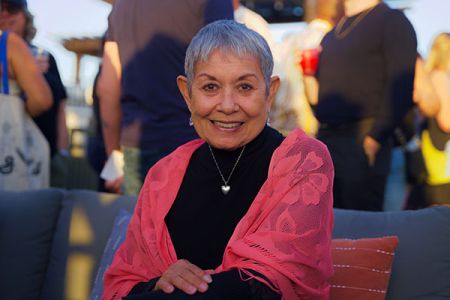 An older woman sitting on an outdoor couch, smiling warmly while wearing a pink shawl.