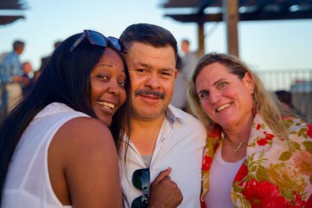 Three people smiling together outdoors at a social event, standing close and enjoying the evening.
