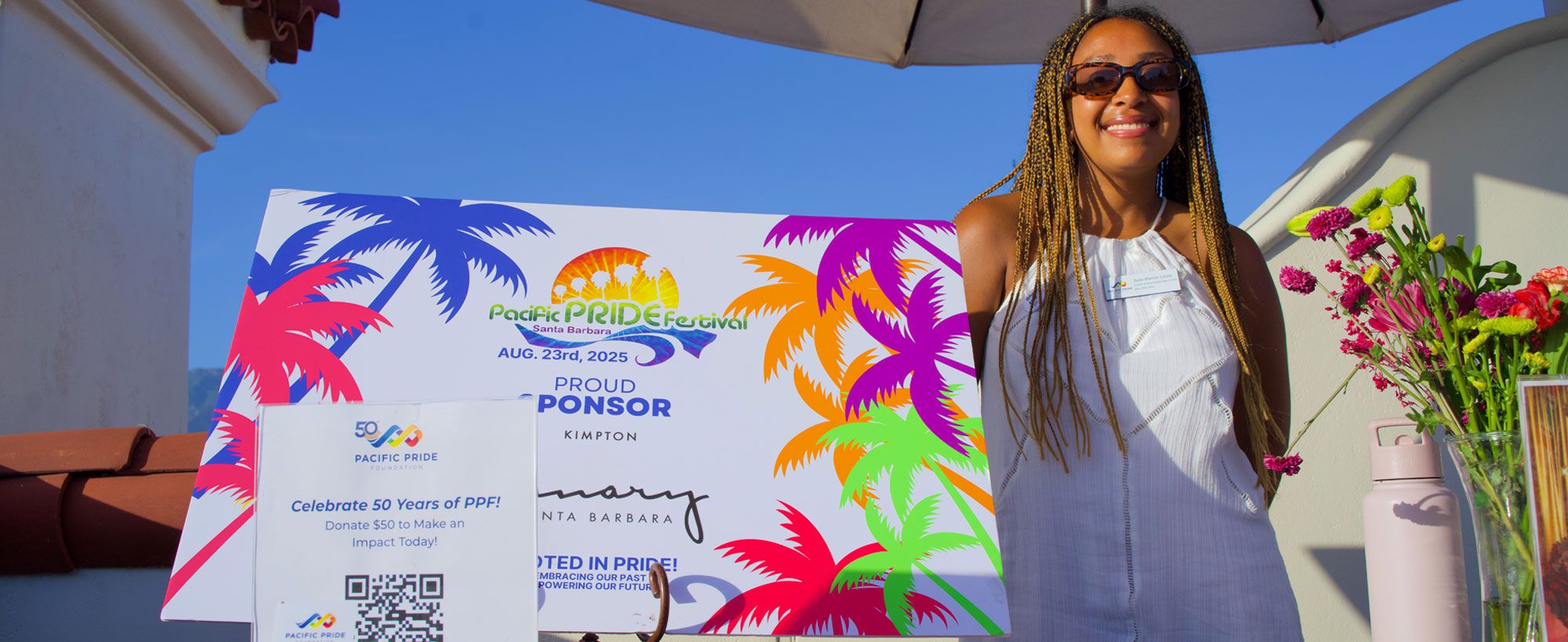 A woman standing beside colorful event signage for the Pacific Pride Festival Santa Barbara 2025, with flowers and a donation display on the table.