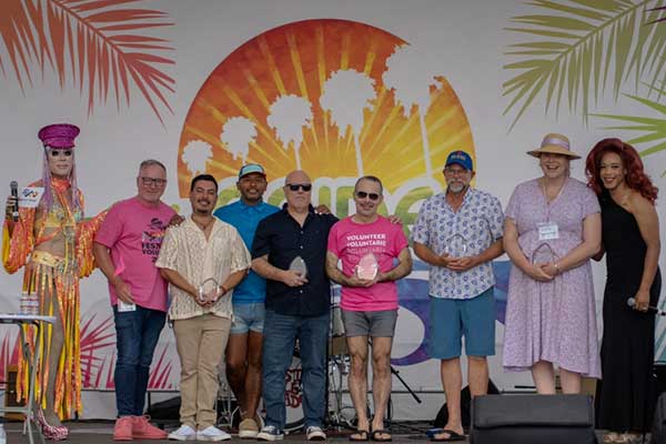 A group of ten people stand together onstage at the Pacific Pride Festival after receiving Community Pride Champion Awards, smiling in front of a bright rainbow and palm tree backdrop.