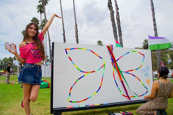 A young woman poses next to a large interactive butterfly wing mural at the Pacific Pride Festival, while another person sits nearby coloring it in with rainbow tape.