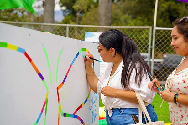 Two attendees color in a butterfly wing mural at the Pacific Pride Festival, using markers and rainbow-colored tape on a whiteboard labeled “Color Me.”