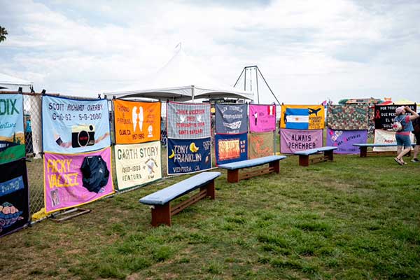 Colorful memorial quilts hang along a fence at the Pacific Pride Festival, each honoring loved ones lost, with benches placed in front for reflection.