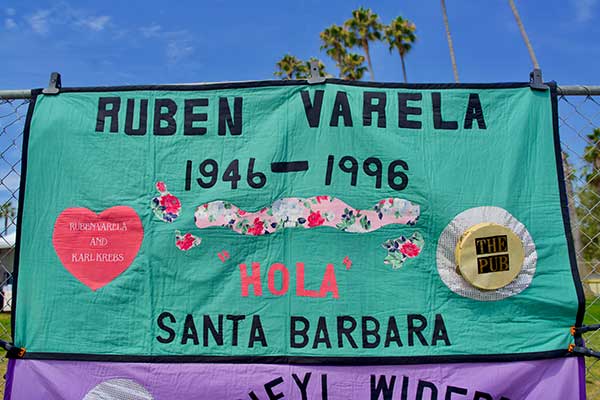 Close-up of a quilt panel honoring Ruben Varela, 1946–1996, from Santa Barbara, decorated with roses, the word “Hola,” and fabric patches referencing “The Pub” and “Ruben Varela and Karl Krebs.