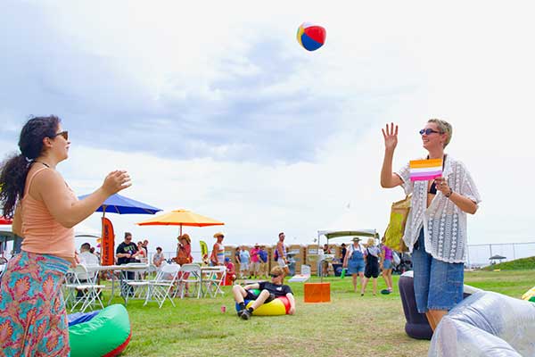 Attendees enjoy the Family Zone at the Pacific Pride Festival, playing with a beach ball and relaxing under colorful umbrellas on the grass.
