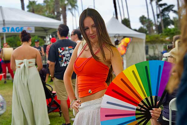 A smiling woman poses at the Pacific Pride Festival wearing a red crop top and white skirt, while someone next to her holds a large rainbow-colored fan.