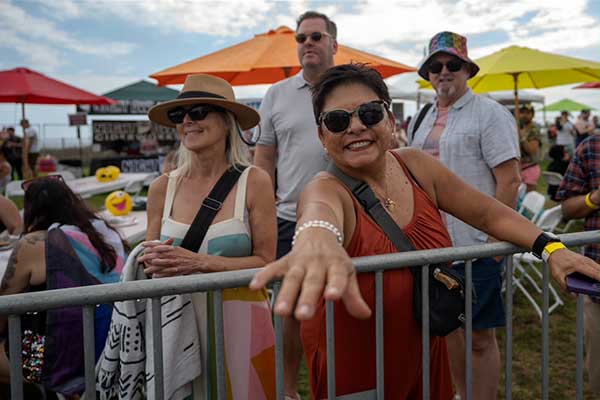 Festivalgoers stand behind a metal barricade at the Pacific Pride Festival, smiling and arm stretched out toward the camera under colorful umbrellas.