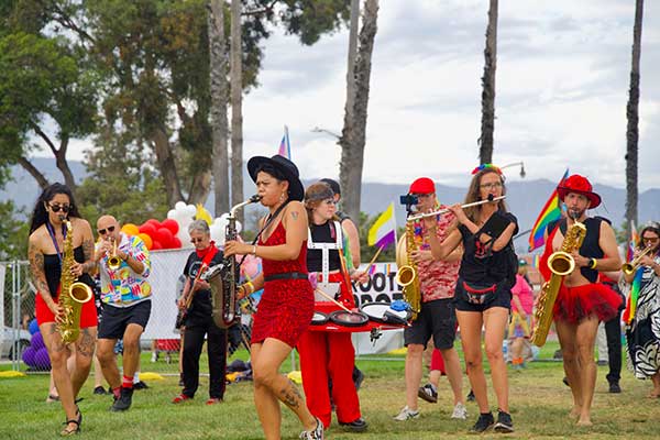 A lively marching band performs at the Pacific Pride Festival, with musicians playing saxophones and flutes while walking across the lawn, surrounded by pride flags and balloons.