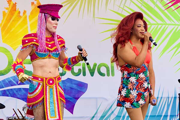 Two performers host onstage at the Pacific Pride Festival; one wears a bright, sequined costume with a headdress, and the other wears a tropical floral dress, both holding microphones in front of a colorful festival backdrop.