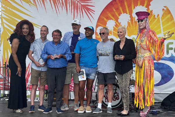 A group of eight people, including two hosts, pose onstage at the Pacific Pride Festival after receiving Pride Legacy Awards, standing in front of a colorful backdrop with palm fronds and a rainbow sunburst.